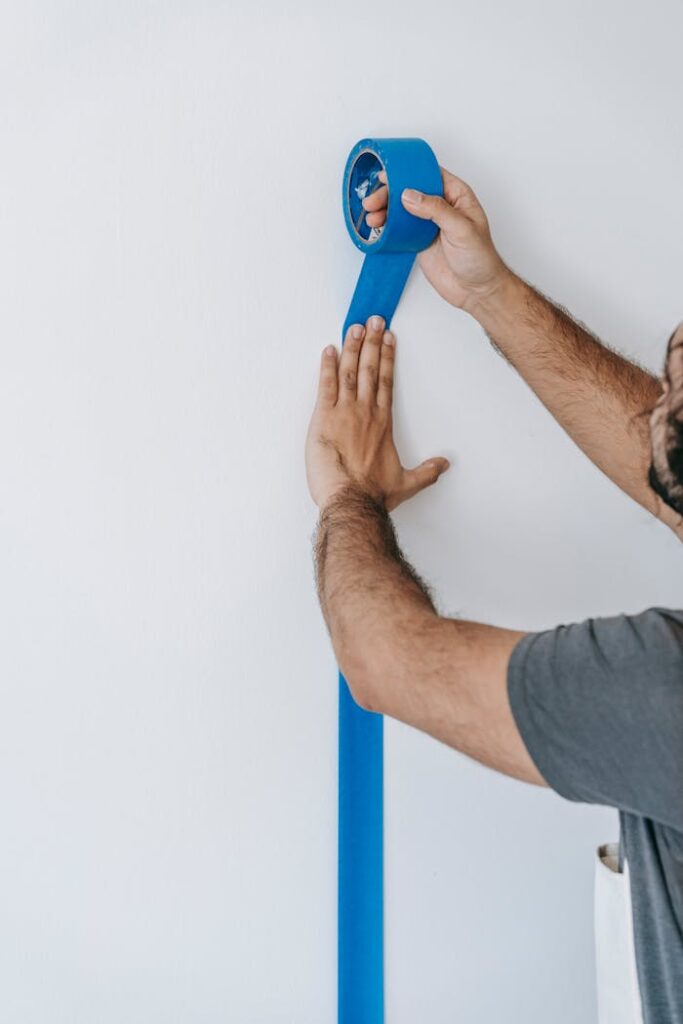 Close-up of a person applying blue tape on a white wall, preparing for painting.